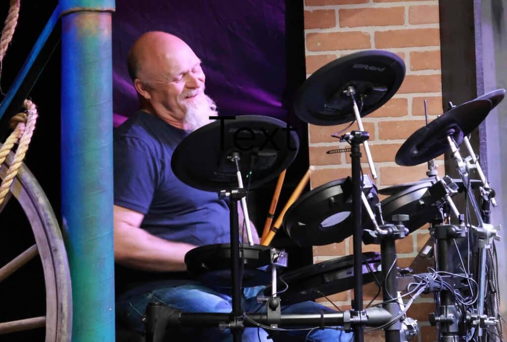 Bald man with a beard sits at an electric drum kit with drumsticks in his hand, laughing
