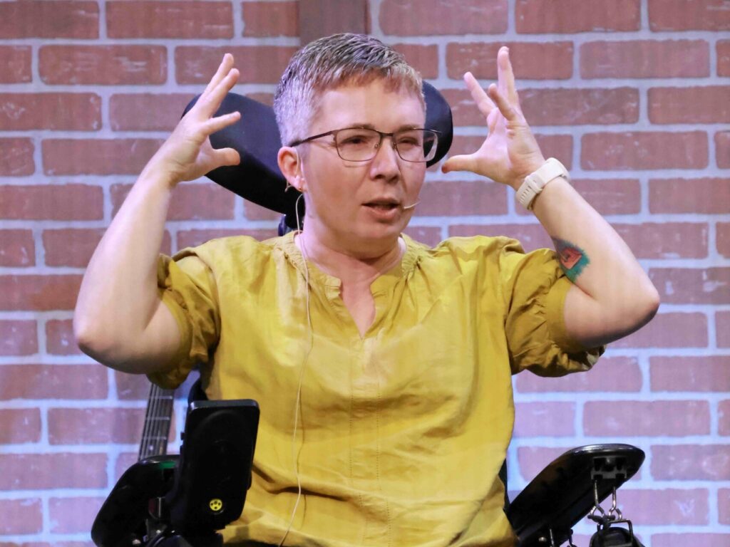 Woman sits in a power wheelchair speaking, gesturing with her hands up by her head